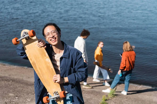 Joyeux asiatique homme avec longboard regarder caméra près de rivière et flous amis — Photo de stock
