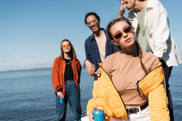 Joven mujer en gafas de sol sosteniendo soda lata y mirando a la cámara cerca borrosa interracial amigos - foto de stock