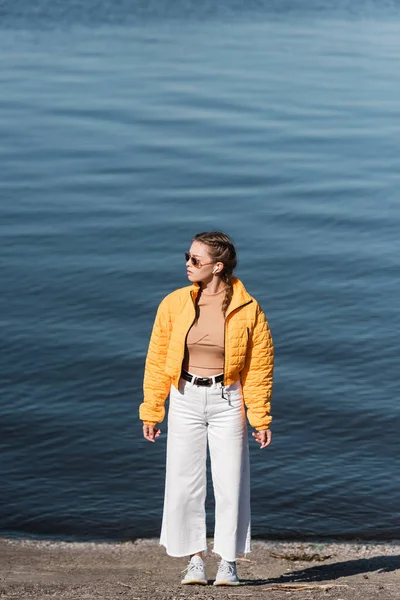 Pleine longueur de femme en pantalon blanc et veste jaune regardant loin sur la rive de la rivière — Photo de stock