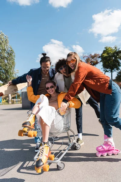 Mulher em rolos patins acenando mão no carrinho de compras perto de amigos multiétnicos alegres — Fotografia de Stock