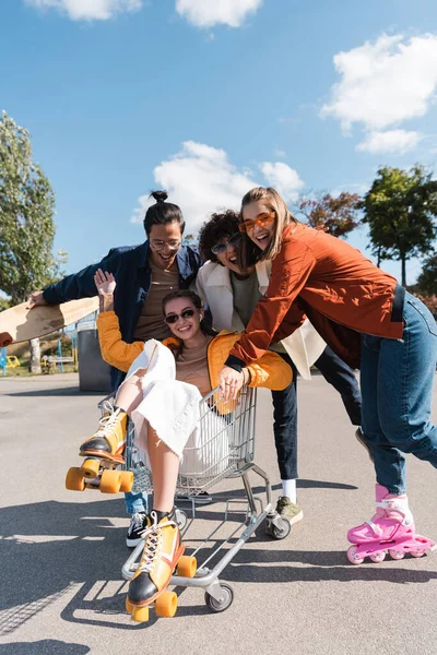 Woman in rollers skates waving hand in shopping cart near cheerful multiethnic friends — Stock Photo