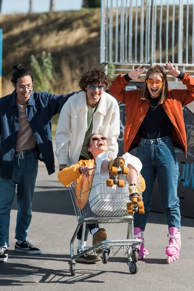 Mujer emocionada riendo en carrito de compras cerca de amigos multiétnicos al aire libre - foto de stock