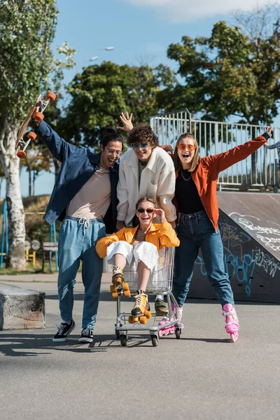 Excited multiethnic skaters moving shopping cart with happy woman in skate park — Stock Photo