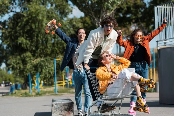 Jóvenes y alegres patinadores multiétnicos se divierten con carrito de compras en el parque - foto de stock