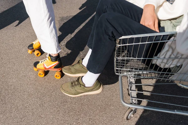 Partial view of man in shopping cart and woman on rollers skates outdoors — Stock Photo