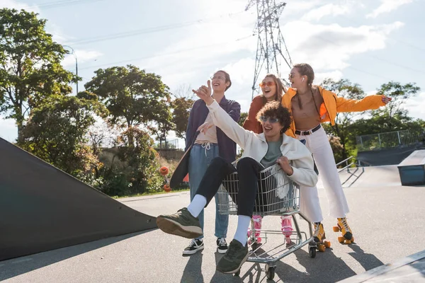 Joyful man showing shaka gesture while riding in shopping trolley near multiethnic friends — Stock Photo