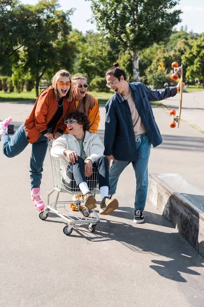 Happy skaters moving shopping trolley with friend while having fun in park — Stock Photo