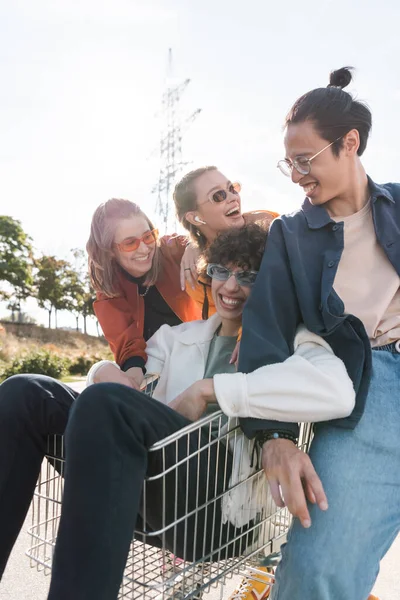 Multiethnic friends laughing near man in shopping trolley — Stock Photo