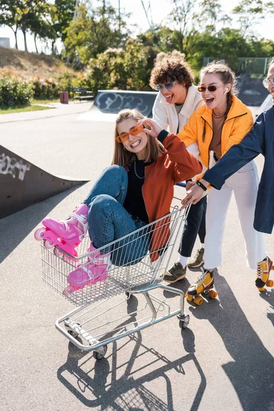 Mulher sorrindo ajustando óculos de sol enquanto montava no carrinho de compras perto de amigos — Fotografia de Stock