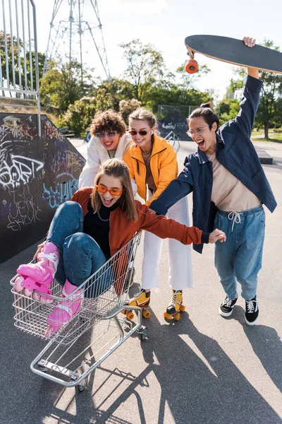 Excitadas mujeres gritando en carrito de compras cerca de amigos interracial divirtiéndose en skate park - foto de stock