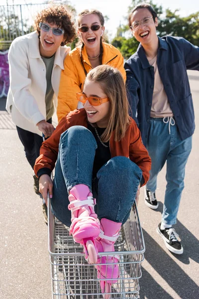 Alegre multicultural amigos moviendo carrito de compras con alegre mujer en gafas de sol - foto de stock
