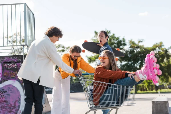 Mulher alegre em rolos de skate sentado no carrinho de compras perto animado e chocado amigos multiétnicos — Fotografia de Stock