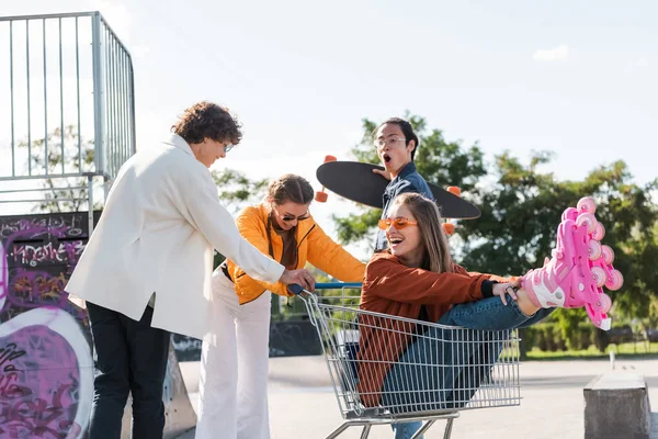 Donna allegra in pattini rotelle seduto nel carrello della spesa vicino eccitato e scioccato amici multietnici — Foto stock