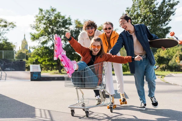 Aufgeregte Frau auf Rollschuhen reitet in Einkaufswagen neben multiethnischen Freunden — Stockfoto