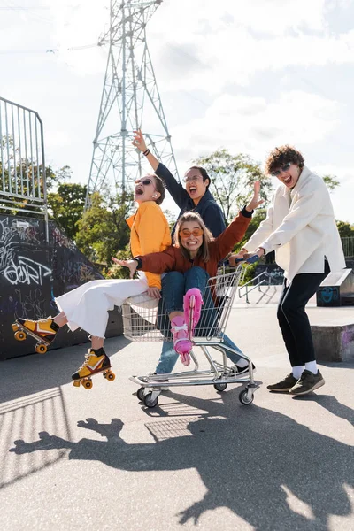 Astonished multicultural friends shouting while having fun with shopping trolley in skate park — Stock Photo