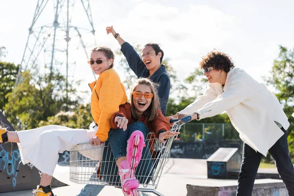 Alegre interracial amigos moviendo carrito de compras con emocionado mujeres al aire libre - foto de stock