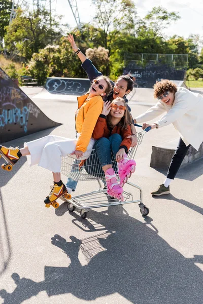 Excited multiethnic friends shouting while riding in shopping cart in skate park — Stock Photo