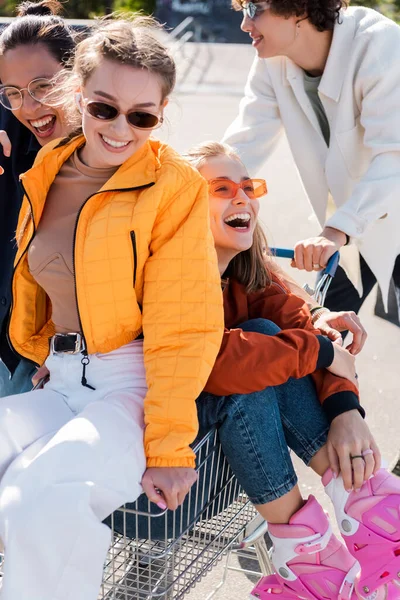 Happy multicultural friends in sunglasses having fun with shopping trolley outdoors — Stock Photo