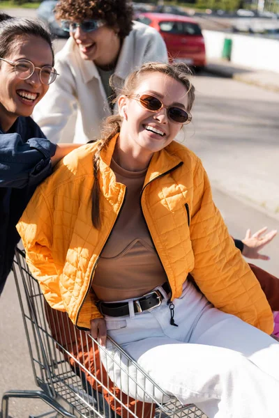 Happy woman in sunglasses sitting in shopping cart near interracial friends — Stock Photo