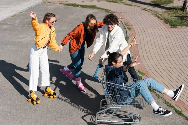 Fröhliche Frauen Händchen haltend und Rollschuhfahren neben asiatischem Mann im Einkaufswagen — Stockfoto