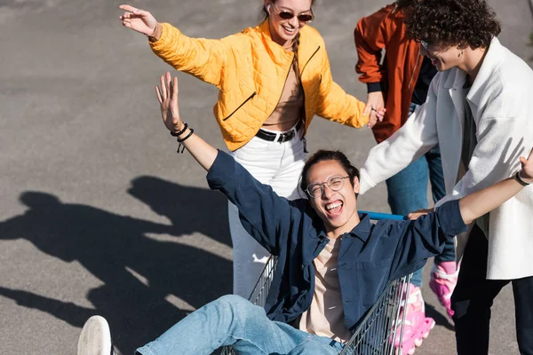 Excited asian man with outstretched hands screaming while riding in shopping cart near friends — Stock Photo