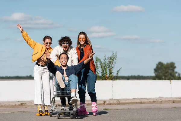 Excited man shouting while moving shopping cart with asian friend near women on rollers skates — Stock Photo