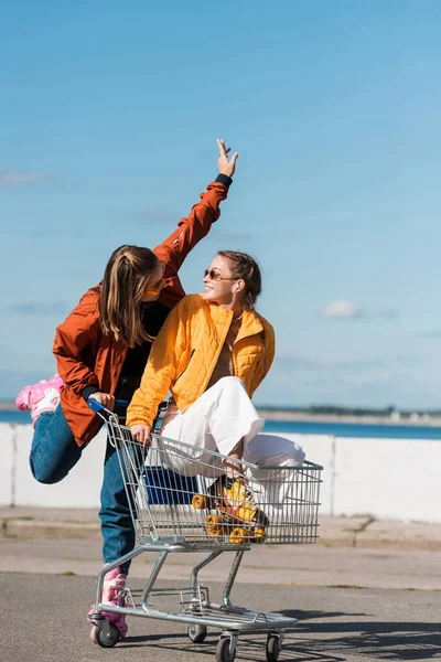 Mujeres felices en patines patines mirándose mientras se divierten con el carrito de compras - foto de stock