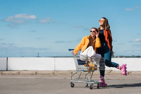 Happy woman in shopping trolley adjusting sunglasses near smiling friend on roller skates — Stock Photo