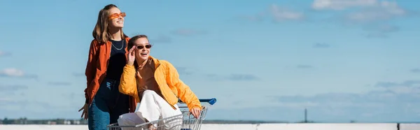 Mulher rindo ajustando óculos de sol enquanto sentado no carrinho de compras perto de amigo, banner — Fotografia de Stock