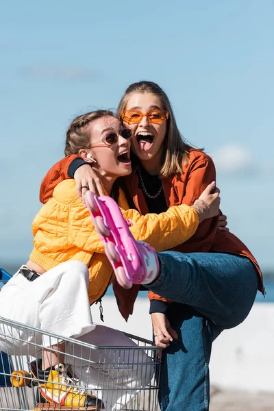 Astonished woman in roller skates embracing friend sitting in shopping cart — Stock Photo