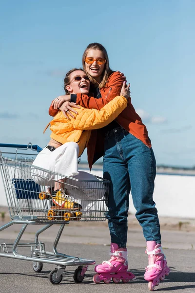 Cheerful woman on roller skates embracing friend sitting in shopping trolley outdoors — Stock Photo