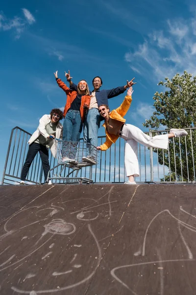 Vista angolo basso di allegri amici multiculturali con carrello della spesa divertirsi sulla rampa di skate — Foto stock