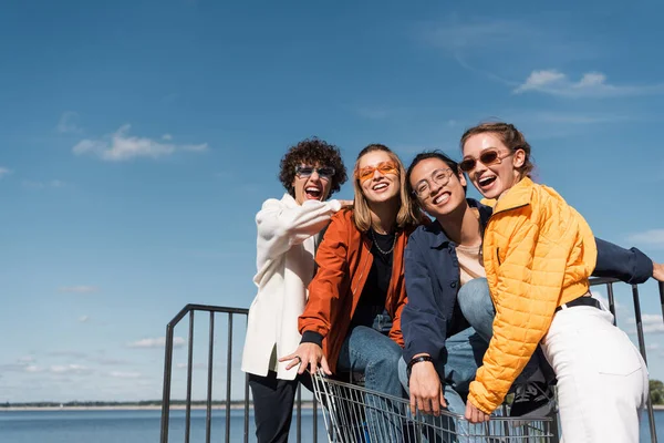 Excited multicultural friends with shopping trolley laughing at camera against blue sky — Stock Photo