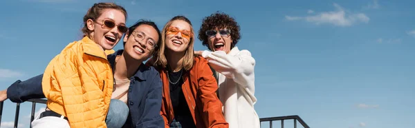 Amigos multiculturales alegres en gafas de sol elegantes mirando a la cámara al aire libre, pancarta - foto de stock