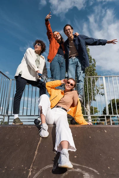 Femme élégante dans des lunettes de soleil près d'amis multiethniques sur la rampe dans le skate park — Photo de stock