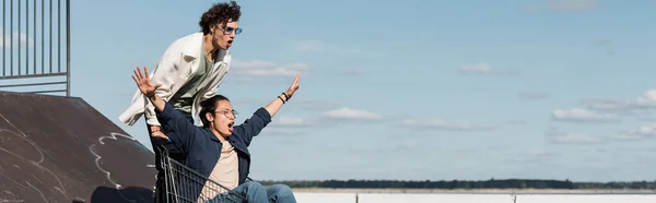 Excited asian man with outstretched hands screaming in shopping cart near friend, banner — Stock Photo