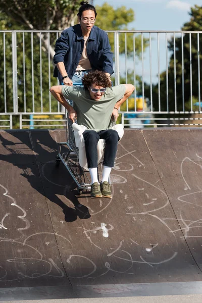 Cheerful interracial friends having fun with shopping cart on ramp in skate park — Stock Photo