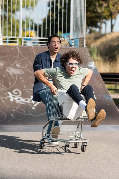 Excité homme hurlant dans shopping trolley près asiatique homme à l'extérieur — Photo de stock