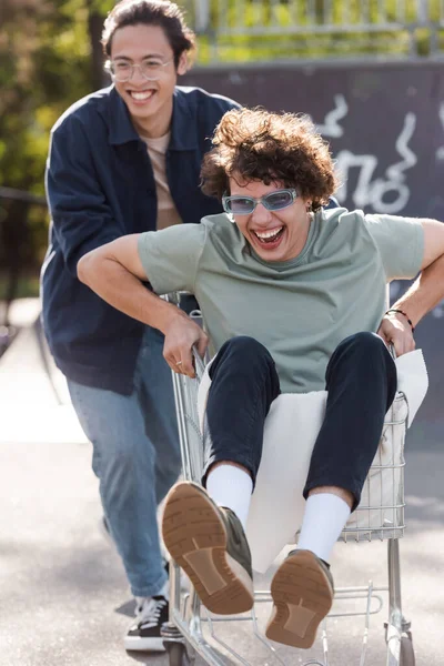 Verschwommener asiatischer Mann bewegt Einkaufswagen mit lachendem Freund im Freien — Stockfoto