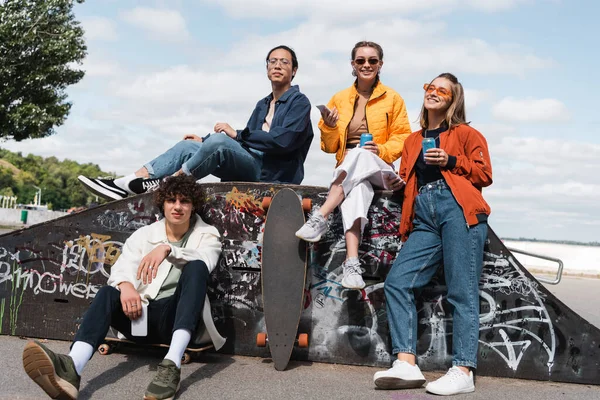 Joyeux patineurs multiethniques avec smartphones et canettes de soda dans le skate park — Photo de stock