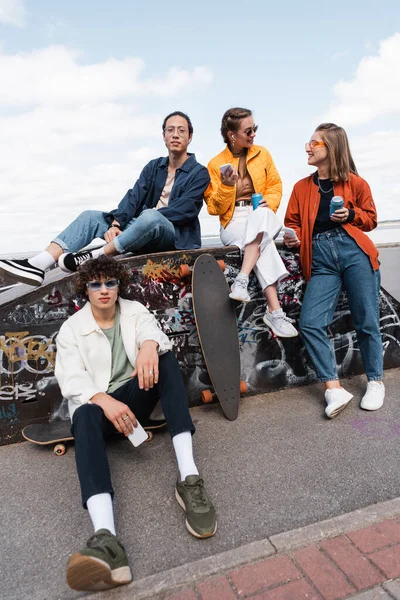 Young women with soda cans sitting on ledge in skate park near interracial skaters — Stock Photo