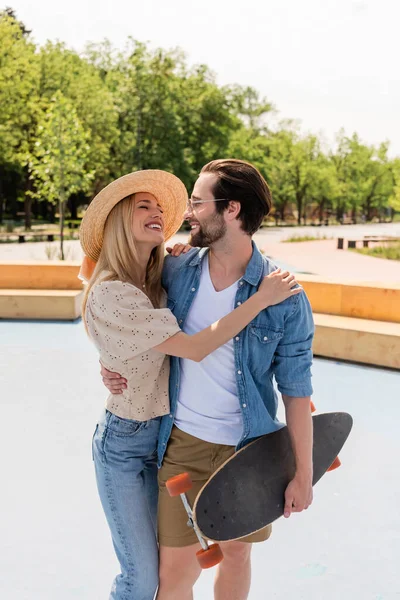 Woman in straw hat hugging boyfriend with longboard in skate park — Stock Photo