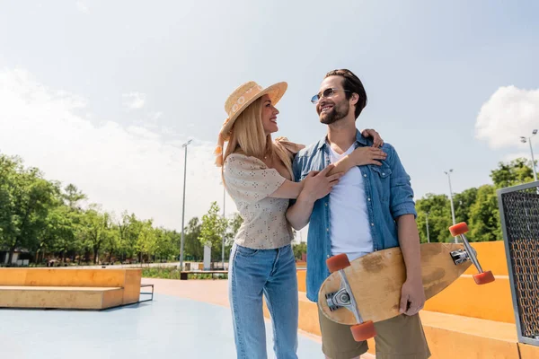 Улыбающаяся женщина в солнцезащитной шляпе обнимает бойфренда держа длинную доску в скейт-парке — стоковое фото
