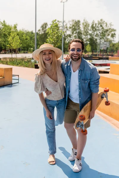 Cheerful couple with longboard hugging and looking at camera in skate park — Stock Photo