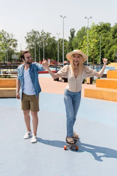Mujer excitada en sombrero de paja montando longboard y cogida de la mano del novio en skate park - foto de stock