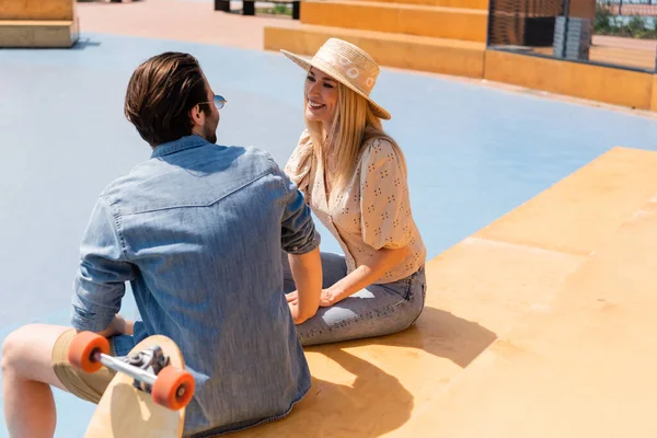 Donna sorridente in cappello da sole che parla con il fidanzato vicino al longboard sfocato nello skate park — Foto stock