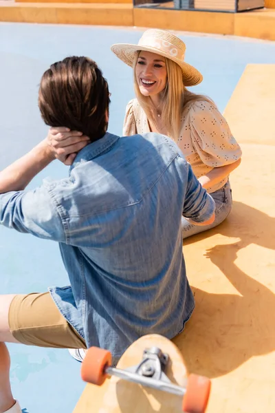Lächelnde Frau mit Strohhut plaudert mit Freund neben Longboard im Skatepark — Stockfoto