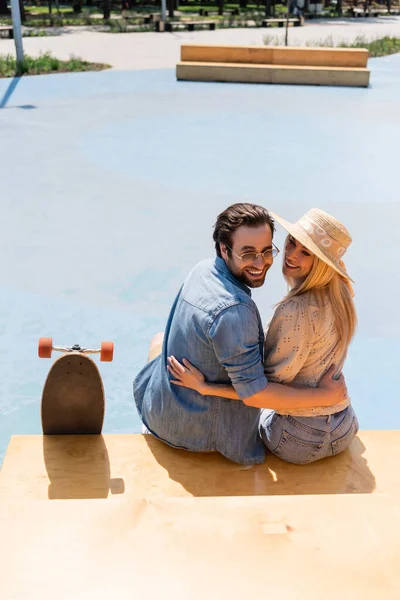 Coppia sorridente in cappello di paglia e occhiali da sole che si abbraccia vicino al longboard nello skate park — Foto stock