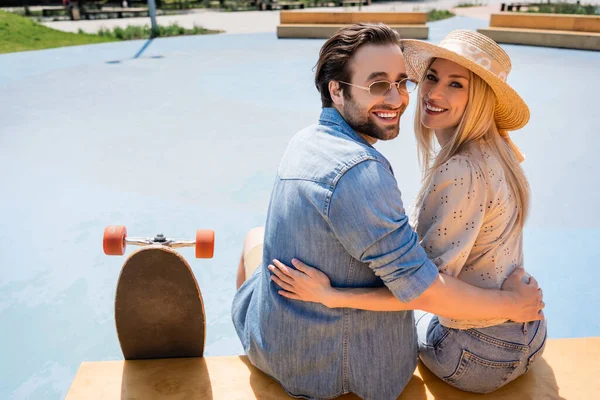 Couple positif étreignant et regardant la caméra près de longboard dans le skate park — Photo de stock