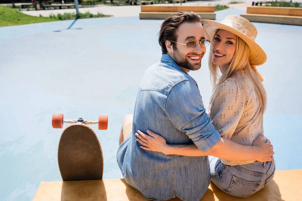 Pareja positiva abrazando y mirando la cámara cerca de longboard en skate park - foto de stock